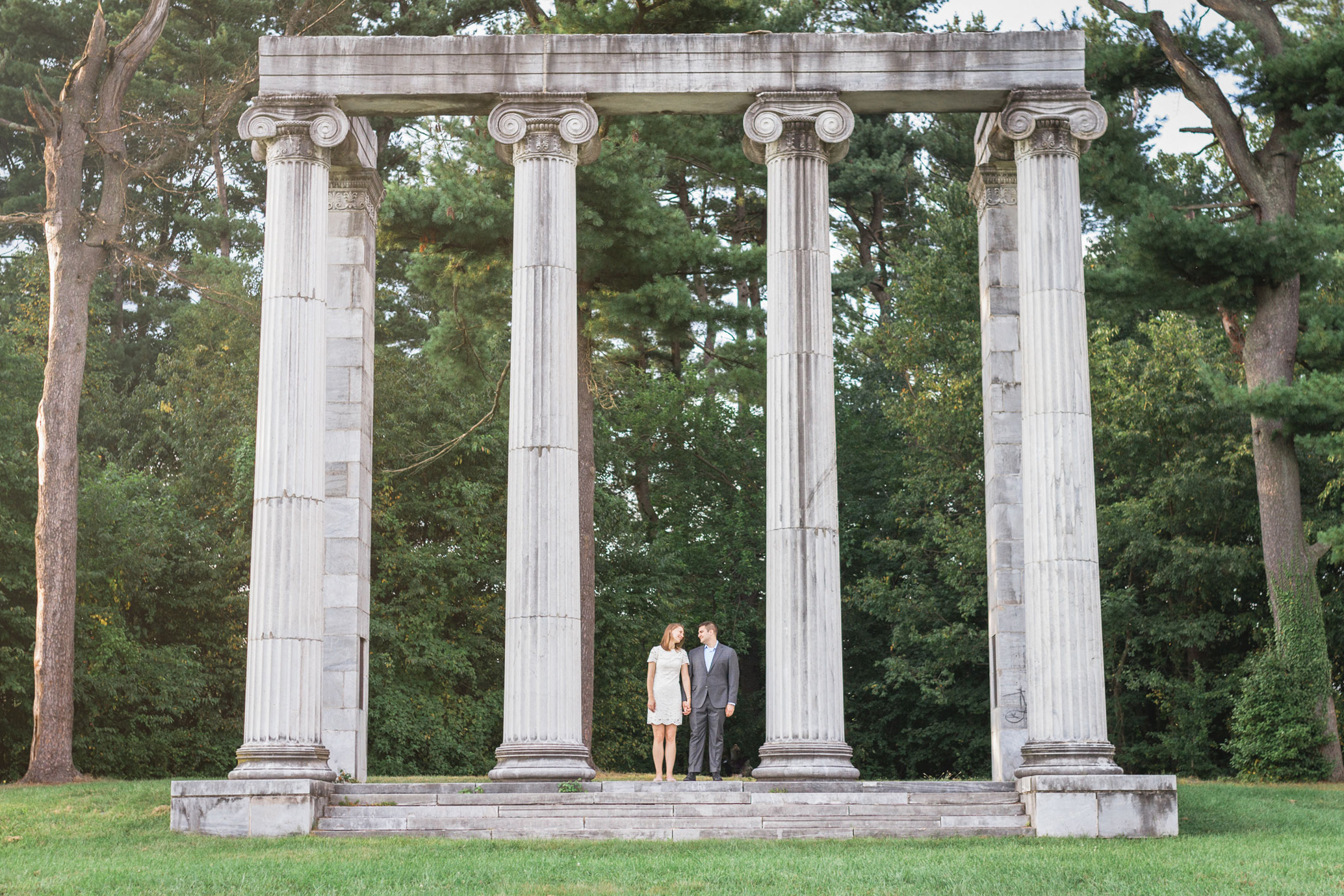 couple standing under Romanesque architecture surrounded by trees